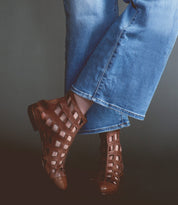 Wearing the Bed|Stü Skyline brown ankle boots handcrafted in Italy, a person in blue jeans stands with legs crossed against a dark background.