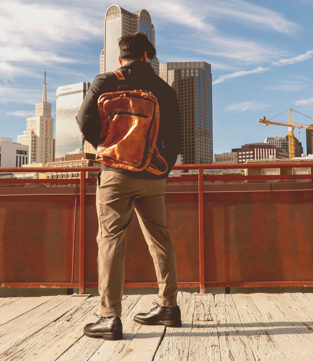 Wearing the Socrates leather backpack by Bed|Stü, a person stands on a wooden walkway, gazing at city buildings and a construction crane beneath a blue sky.