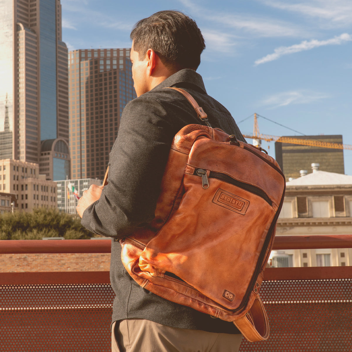 A man stands outdoors in a city with tall buildings in the background, facing away from the camera, wearing the Bed|Stü Socrates brown leather backpack with its crossbody strap subtly visible over his shoulder.