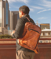 A man stands outdoors in a city with tall buildings in the background, facing away from the camera, wearing the Bed|Stü Socrates brown leather backpack with its crossbody strap subtly visible over his shoulder.