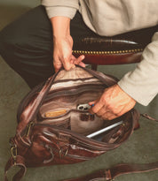 A person sits on a chair and reaches into an open Bed|Stü Socrates brown leather briefcase, revealing a wallet, phone, pen, and a laptop inside.