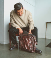 A man sitting on a chair looks down as he places a laptop into the Bed|Stü Socrates brown leather briefcase resting on his lap in a minimalist room.