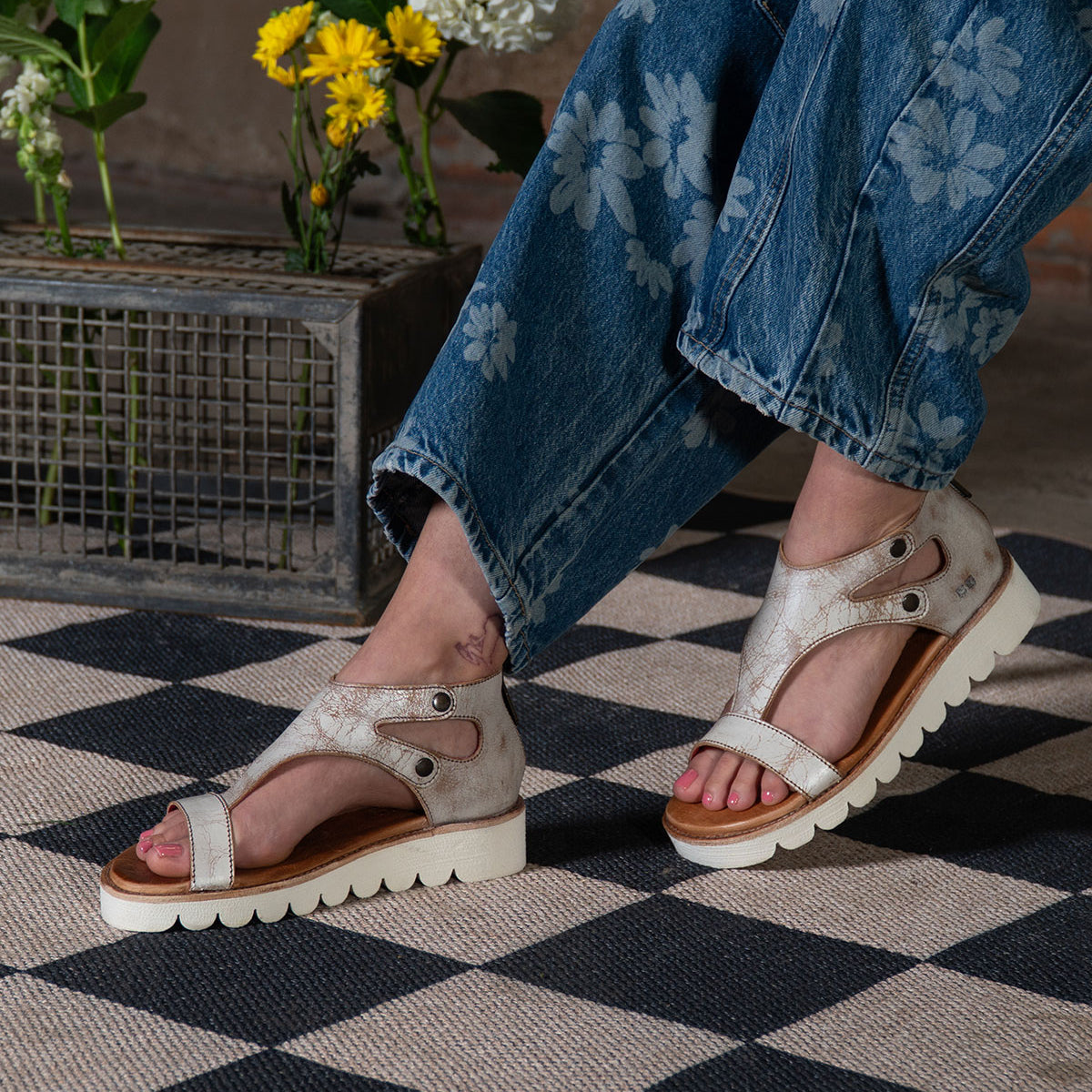 Wearing Bed|Stü's Soni II metallic platform sandals with a durable outsole, this person pairs them with floral-patterned jeans as they sit on a black and white checkered rug near flowers in a metal crate.