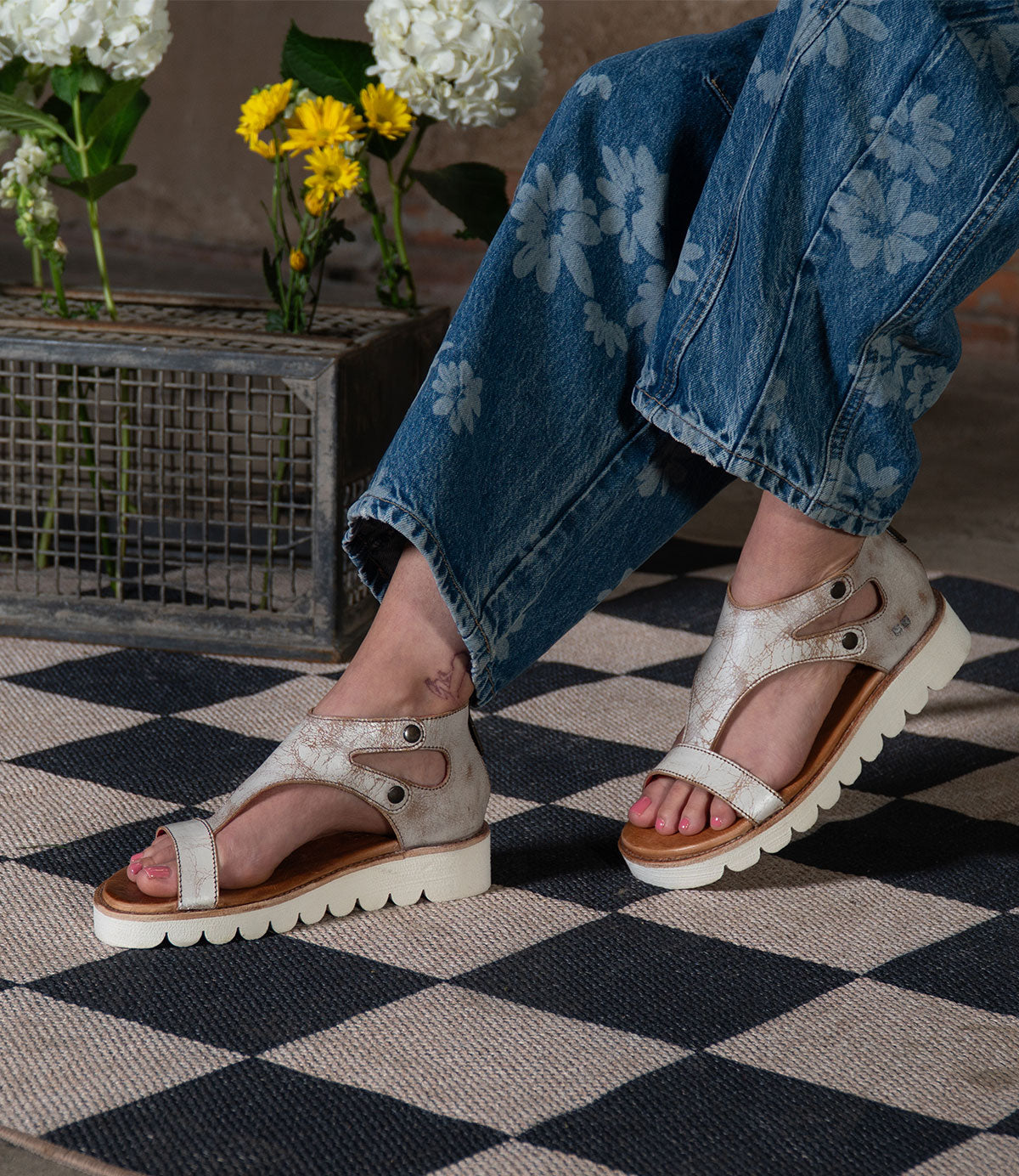 Wearing Bed|Stü's Soni II metallic platform sandals with a durable outsole, this person pairs them with floral-patterned jeans as they sit on a black and white checkered rug near flowers in a metal crate.