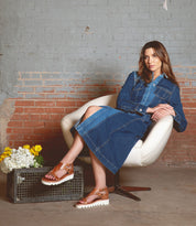 A woman sits in a white chair, wearing a denim dress and the Bed|Stü Soni II brown platform sandals. Nearby, a metal crate filled with white and yellow flowers rests on the floor against a brick wall, complementing the relaxed scene and highlighting her comfortable Soni II sandals with their durable outsole.
