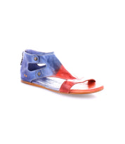 The Bed|Stü Soto is a flat cutout leather sandal featuring an open toe, two ankle buttons, and a striking red and blue color gradient. Designed for everyday exploring, it includes a cushioned footbed for added comfort. Shown on a white background.