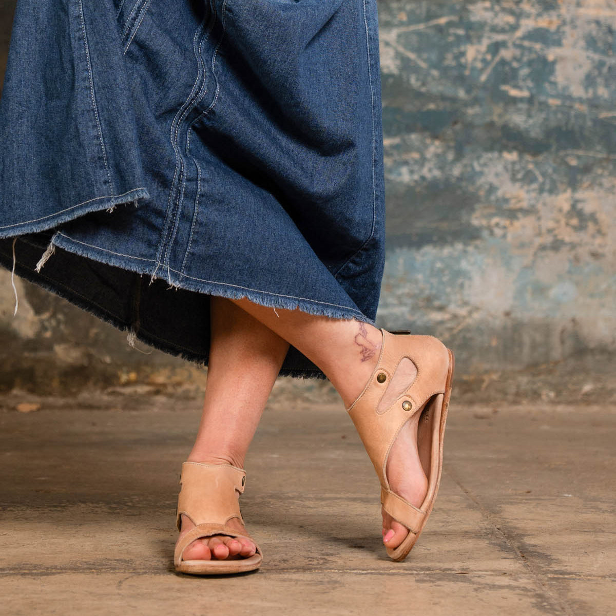 Wearing the Bed|Stü Soto tan cutout leather sandals with a cushioned footbed and a frayed denim skirt, this person stands on a concrete floor, one foot raised to reveal a tattoo on their left ankle—an effortlessly stylish look for everyday adventures.