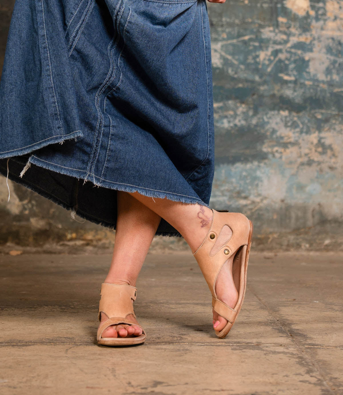 Wearing the Bed|Stü Soto tan cutout leather sandals with a cushioned footbed and a frayed denim skirt, this person stands on a concrete floor, one foot raised to reveal a tattoo on their left ankle—an effortlessly stylish look for everyday adventures.