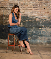 A woman in a denim halter top and jeans sits on a wooden stool against a distressed brick and painted wall, smiling and looking down. She wears the Soto cutout leather sandals by Bed|Stü, featuring a cushioned footbed that’s perfect for everyday exploring.
