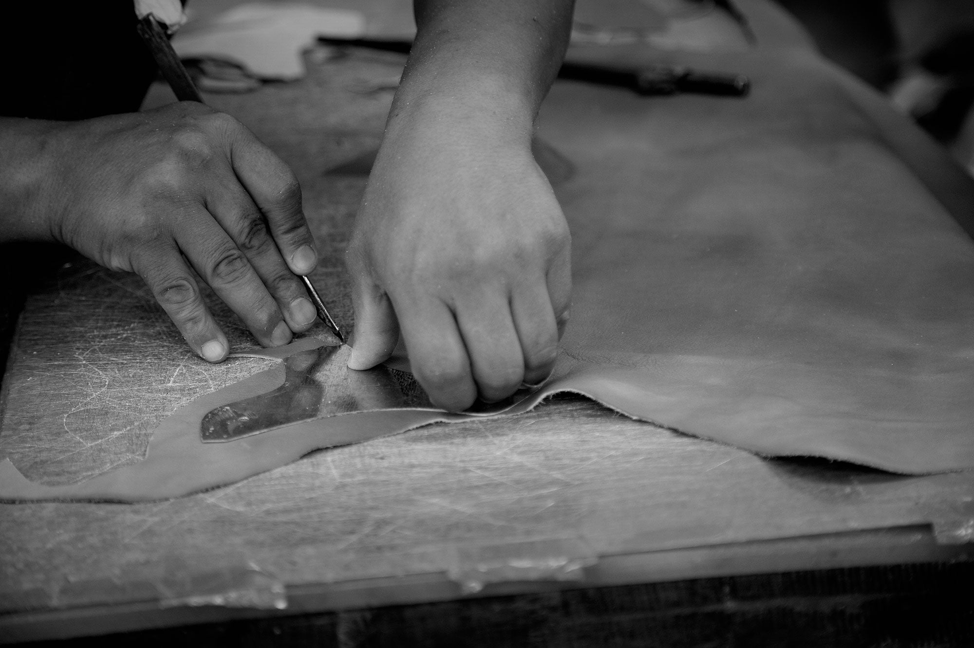 Black-and-white photo of hands cutting leather along a pattern on a worktable.
