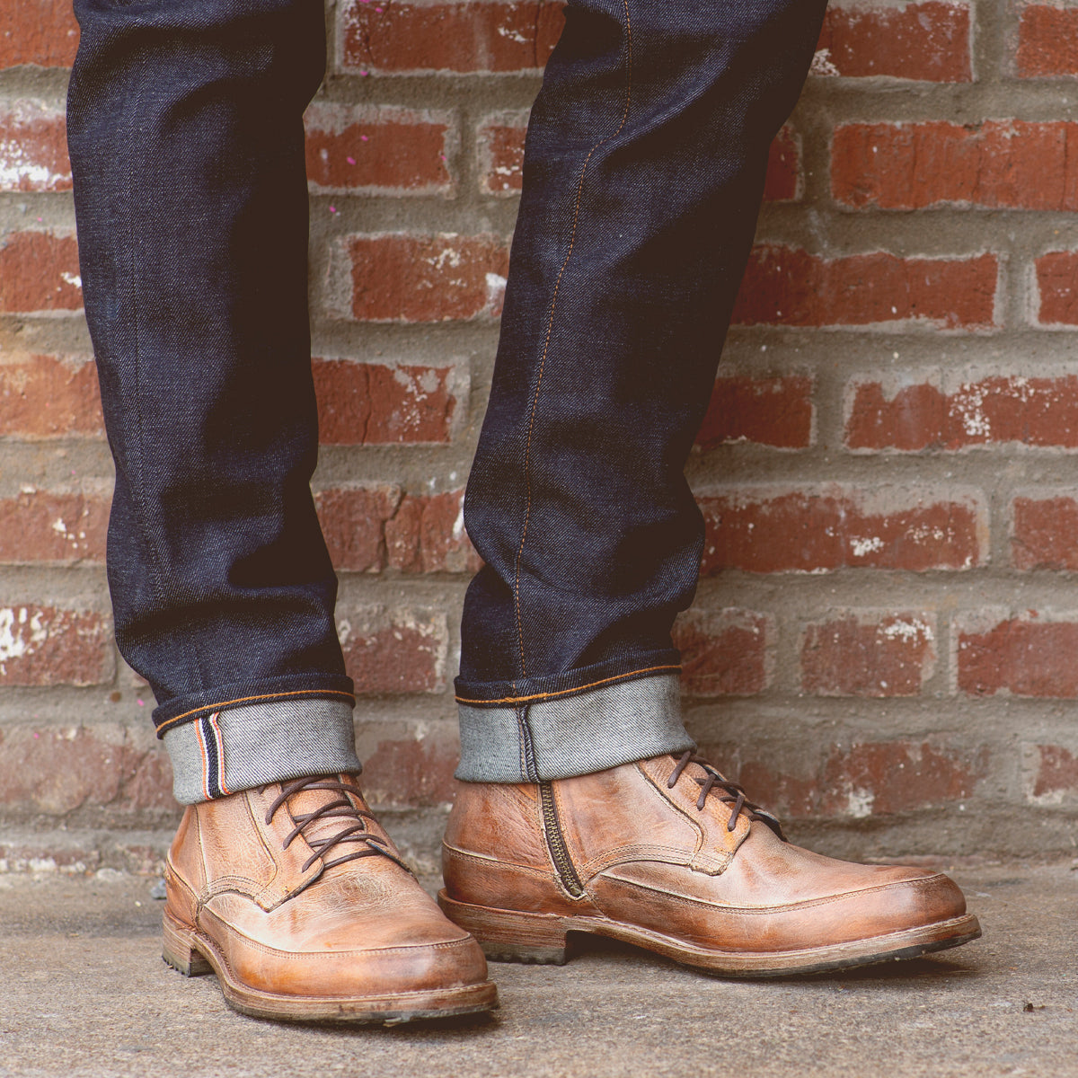Wearing cuffed dark blue jeans, a person stands on a concrete surface in front of a red brick wall, showcasing the Bed|Stü Spiker men’s lace-up boots.