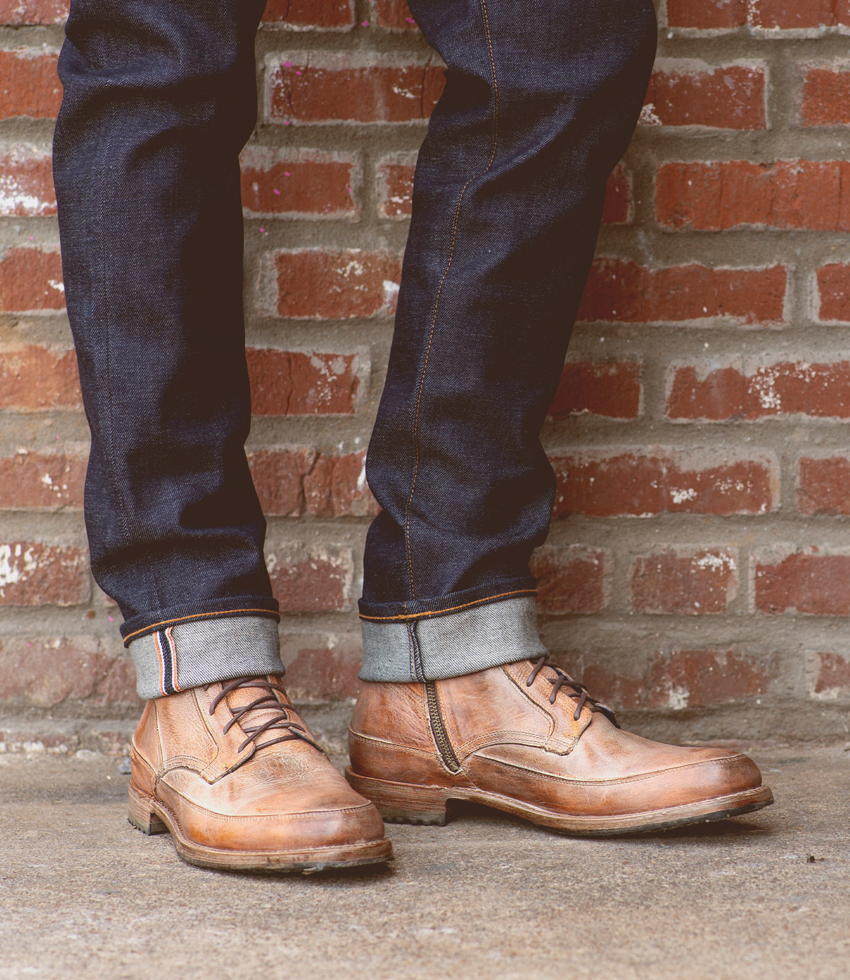 Wearing cuffed dark blue jeans, a person stands on a concrete surface in front of a red brick wall, showcasing the Bed|Stü Spiker men’s lace-up boots.