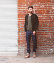 A man stands in front of a brick wall and closed white garage door, wearing a dark cardigan, olive shirt, blue jeans, and the Bed|Stü Spiker rugged men's lace-up boots with a cushioned insole for all-day comfort.