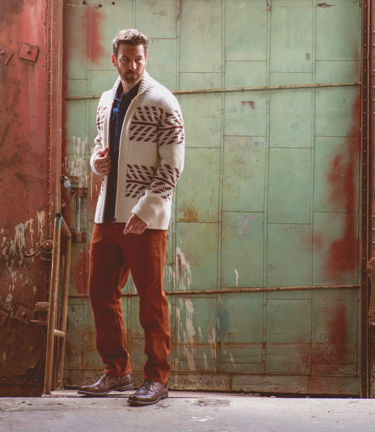 A man wearing a patterned white cardigan, blue shirt, rust-colored pants, and stylish Bed|Stü Spiker men’s lace-up boots stands in front of a large weathered green metal door.