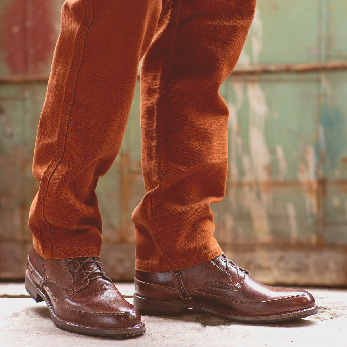 A person wearing Bed|Stü’s Spiker brown leather ankle boots with cushioned insoles and rust-colored pants stands on a concrete surface in front of a weathered green metal door.