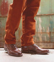 A person wearing Bed|Stü’s Spiker brown leather ankle boots with cushioned insoles and rust-colored pants stands on a concrete surface in front of a weathered green metal door.