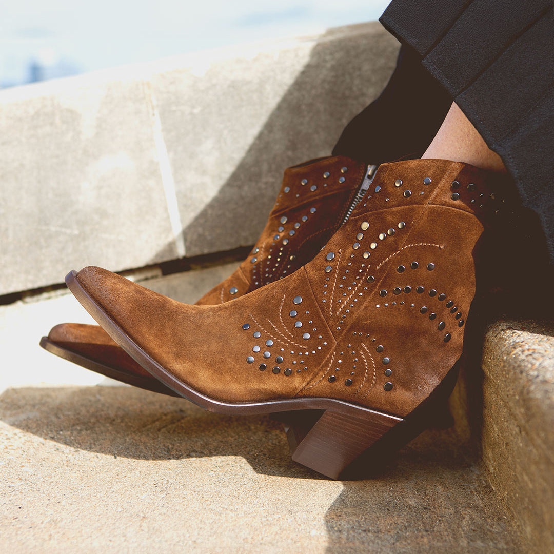 Wearing the Bed|Stü Sposa booties—crafted in brown suede with a pointed toe, decorative stitching, and metal stud accents—paired with black pants and photographed while resting on outdoor stone steps.