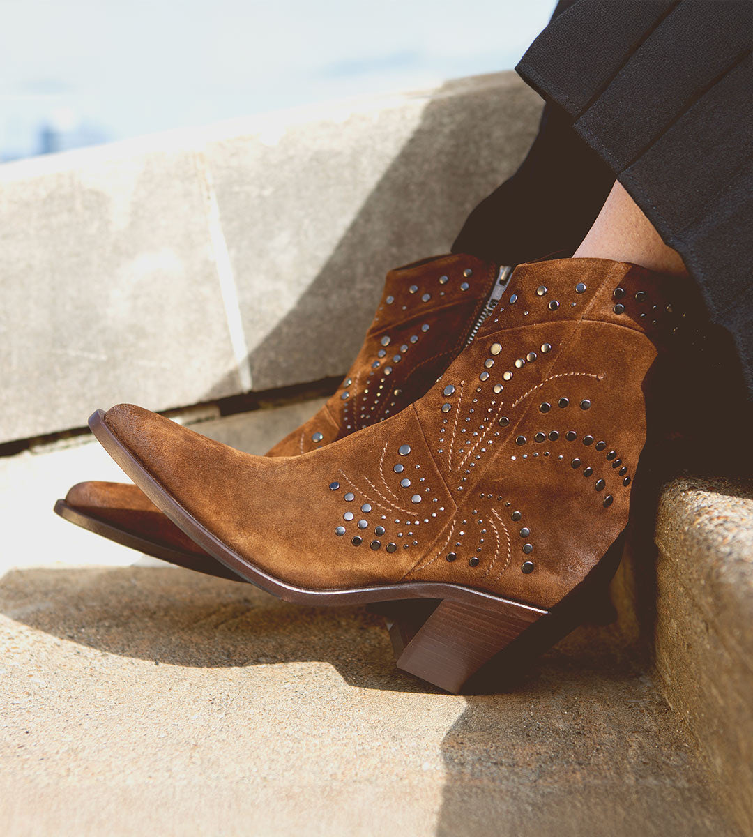 Wearing the Bed|Stü Sposa booties—crafted in brown suede with a pointed toe, decorative stitching, and metal stud accents—paired with black pants and photographed while resting on outdoor stone steps.