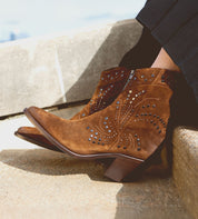Wearing the Bed|Stü Sposa booties—crafted in brown suede with a pointed toe, decorative stitching, and metal stud accents—paired with black pants and photographed while resting on outdoor stone steps.