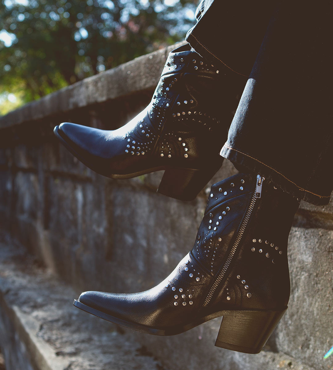 A person wearing the Bed|Stü Sposa black studded cowboy boots and black jeans sits on a stone ledge outdoors, legs hanging over the edge. The pointed toe design of the Sposa booties brings a modern touch to their rugged style.
