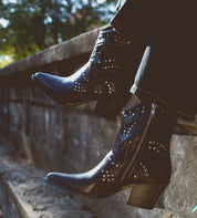 A person wearing the Bed|Stü Sposa black studded cowboy boots and black jeans sits on a stone ledge outdoors, legs hanging over the edge. The pointed toe design of the Sposa booties brings a modern touch to their rugged style.