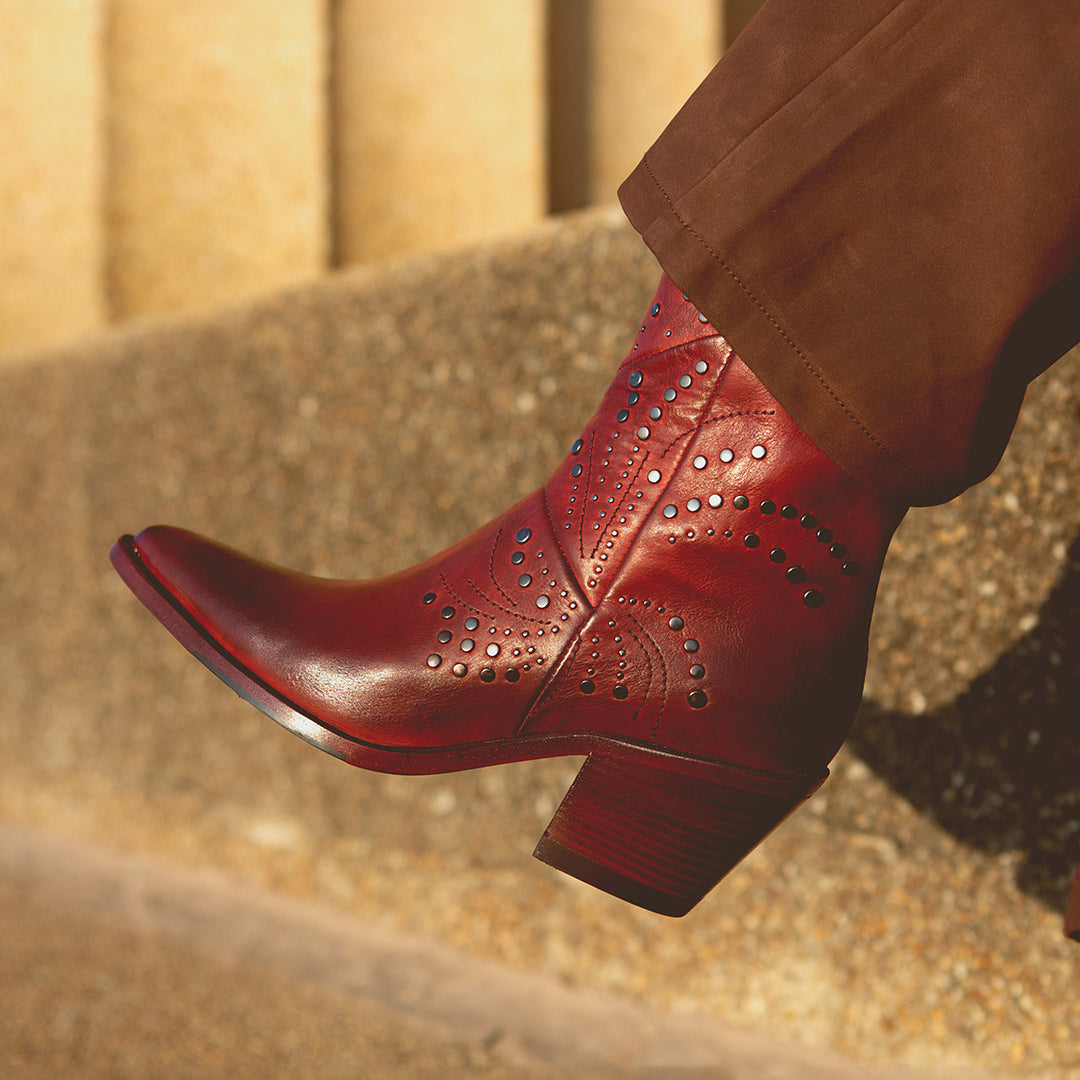 A person wearing brown pants and the Bed|Stü Sposa, an Italian ankle boot crafted from red vegetable-tanned leather with studded patterns, stands on a textured stone surface.