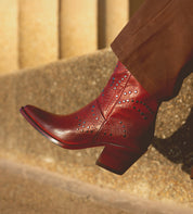 A person wearing brown pants and the Bed|Stü Sposa, an Italian ankle boot crafted from red vegetable-tanned leather with studded patterns, stands on a textured stone surface.