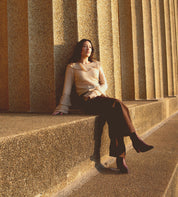 A woman in a beige top and brown pants sits on a ledge against a textured columned wall, with sunlight casting shadows, her look completed by the stylish Sposa pointed toe booties from Bed|Stü, crafted from vegetable-tanned leather.