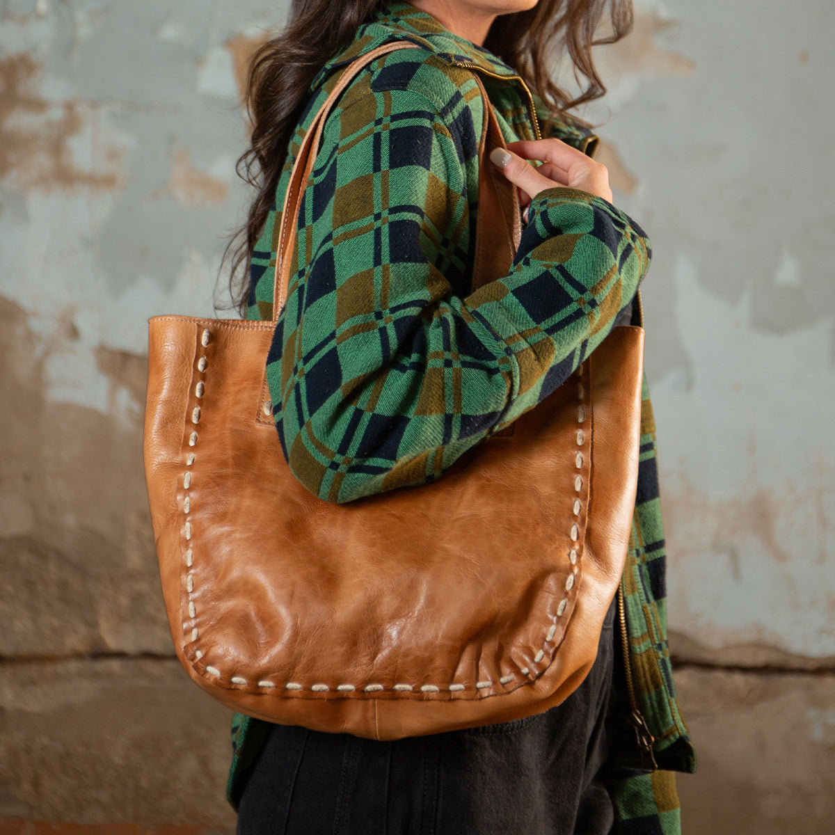 A woman wearing a green plaid shirt stands in front of a textured wall, carrying the Stevie vintage leather tote bag by Bed|Stü over her shoulder.