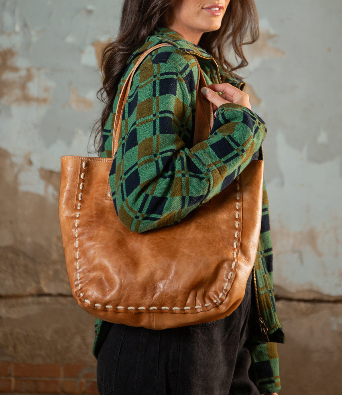 A woman wearing a green plaid shirt stands in front of a textured wall, carrying the Stevie vintage leather tote bag by Bed|Stü over her shoulder.