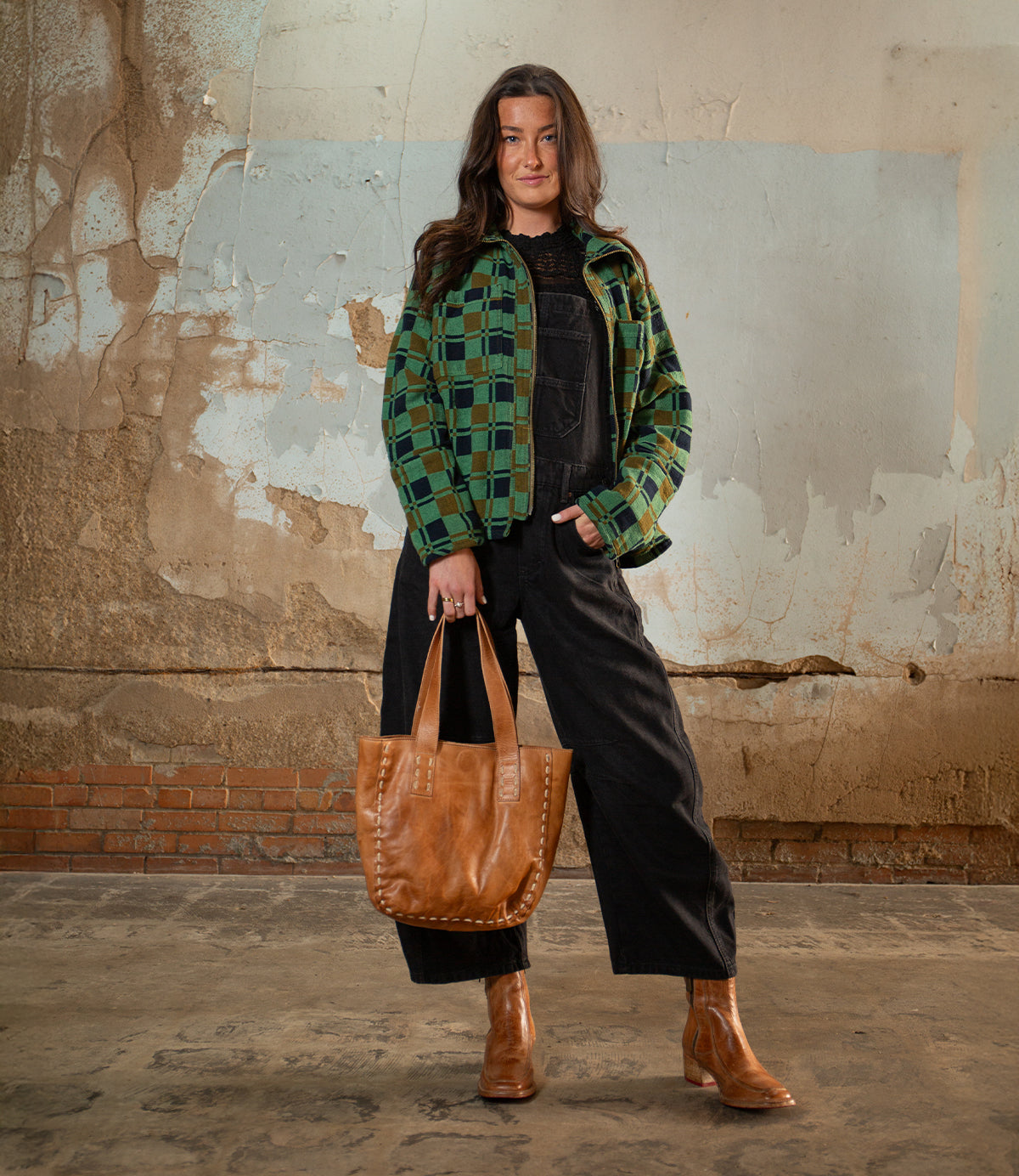 A person stands indoors against a textured wall, wearing a green checkered jacket, black overalls, brown boots, and holding the Stevie tote by Bed|Stü—a vintage leather bag perfect for everyday carry.