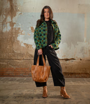 A person stands indoors against a textured wall, wearing a green checkered jacket, black overalls, brown boots, and holding the Stevie tote by Bed|Stü—a vintage leather bag perfect for everyday carry.