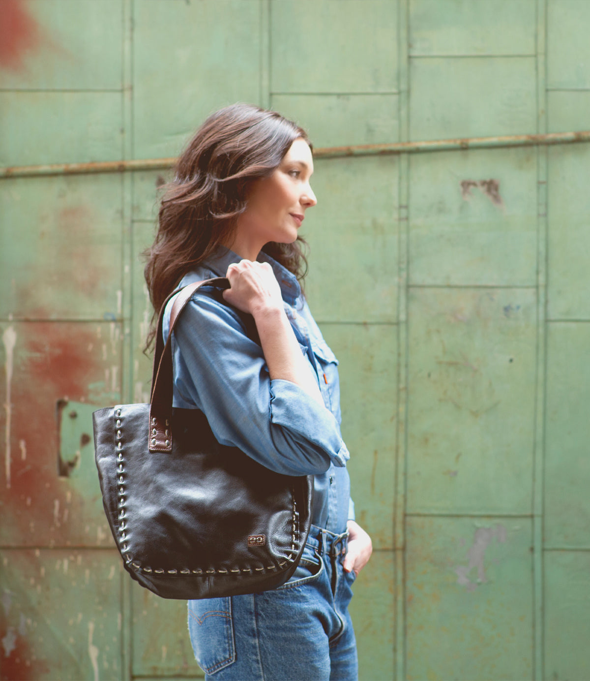 A woman in a denim shirt and jeans stands sideways, holding the Bed|Stü Stevie tote—a sleek black leather bag with visible stitching, perfect for everyday use—set against a weathered green metal background.