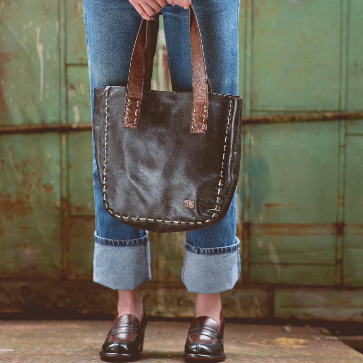 Wearing cuffed jeans and loafers, a person stands before a distressed green metal background, holding the Stevie by Bed|Stü—a large vintage leather tote bag with brown handles that makes for an ideal everyday carry accessory with timeless style.