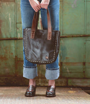 Wearing cuffed jeans and loafers, a person stands before a distressed green metal background, holding the Stevie by Bed|Stü—a large vintage leather tote bag with brown handles that makes for an ideal everyday carry accessory with timeless style.
