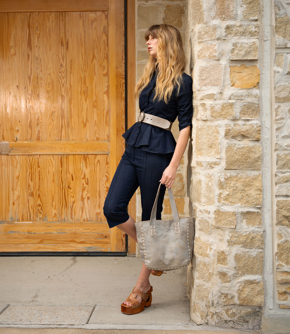 A woman in a navy outfit and beige belt leans against a stone wall, holding the Stevie vintage tote bag by Bed|Stü—perfect for everyday carry. She wears wooden platform sandals and stands near a wooden door.