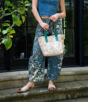 A person in a blue sleeveless top and patterned jeans stands outside holding the Stevie bag by Bed|Stü, wearing tan sandals—making this vintage-inspired accessory the perfect everyday carry.