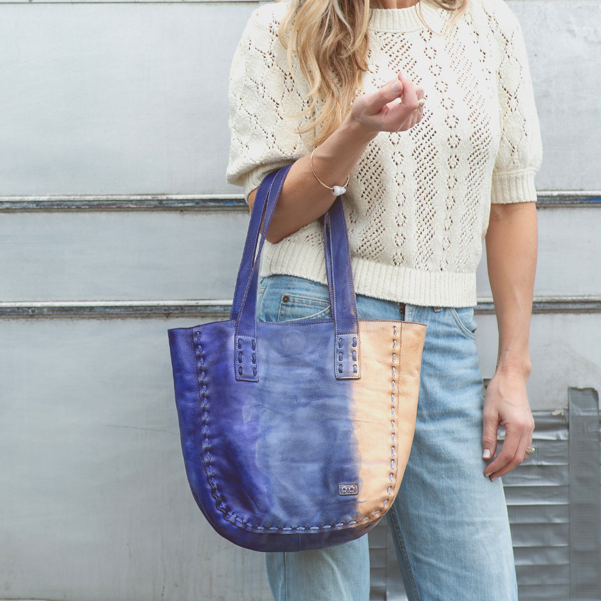 A woman wearing a cream knit sweater and jeans holds the Stevie leather tote by Bed|Stü—an effortlessly stylish bag perfect for everyday carry—as she stands in front of a silver metal wall.