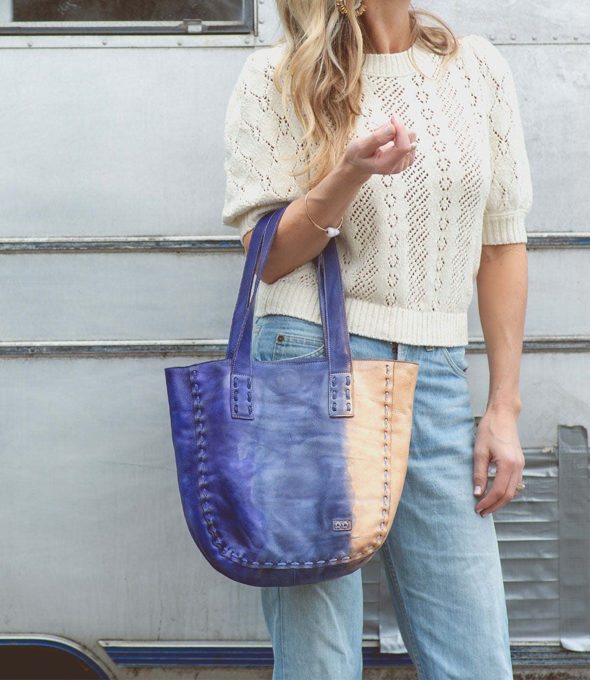 A woman wearing a cream knit sweater and jeans holds the Stevie leather tote by Bed|Stü—an effortlessly stylish bag perfect for everyday carry—as she stands in front of a silver metal wall.