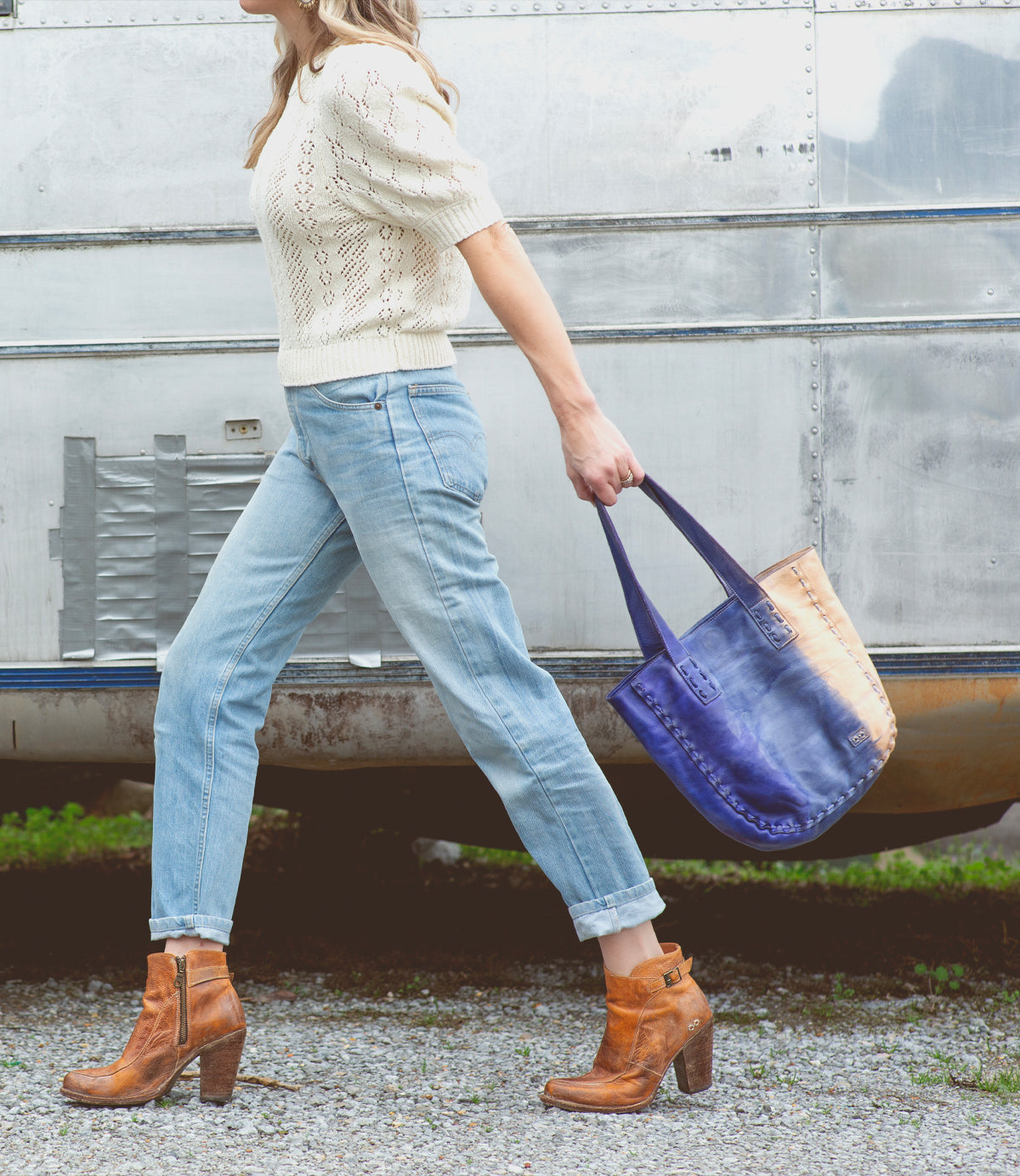 A person walks on gravel in front of a metal trailer, wearing cuffed jeans, a cream sweater, brown heeled boots, and carrying the Stevie leather tote by Bed|Stü as their everyday carry.