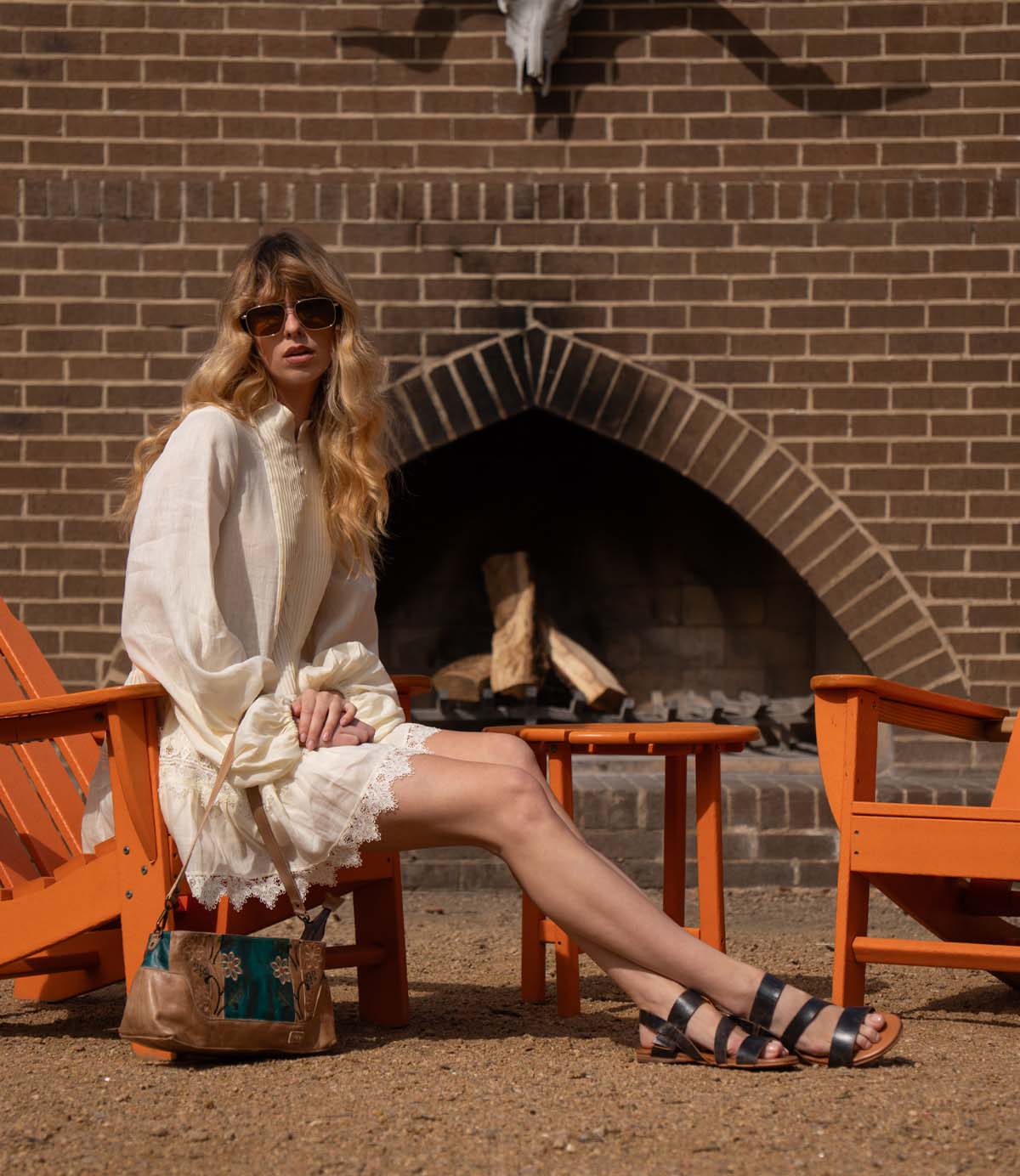 A woman in a white dress and Bed|Stü Superb adjustable ankle strap leather sandals sits on an orange chair by an outdoor fireplace, with a small table and another chair nearby, effortlessly showcasing her timeless style for everyday wear.