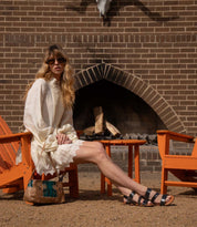 A woman in a white dress and Bed|Stü Superb adjustable ankle strap leather sandals sits on an orange chair by an outdoor fireplace, with a small table and another chair nearby, effortlessly showcasing her timeless style for everyday wear.