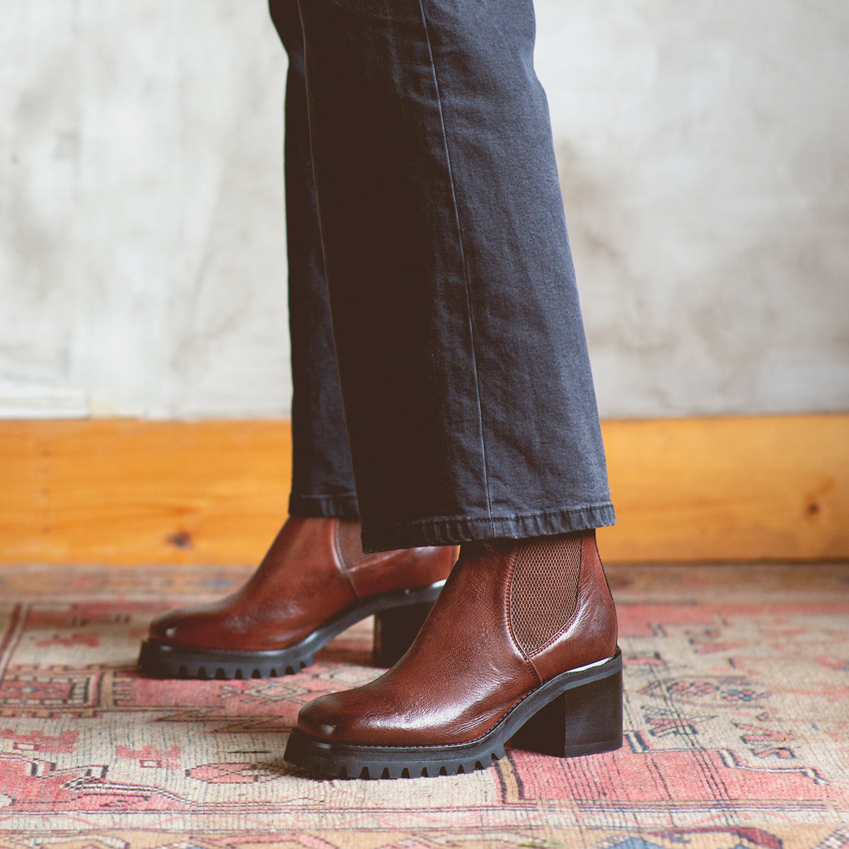 A person wearing dark pants and the Bed|Stü Surge brown Italian leather heeled boots stands on a patterned rug, with a light-colored wall and wooden trim in the background.