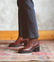 A person wearing dark pants and the Bed|Stü Surge brown Italian leather heeled boots stands on a patterned rug, with a light-colored wall and wooden trim in the background.