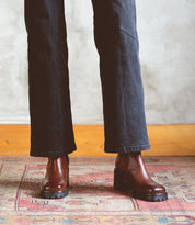 A person wearing dark pants and Bed|Stü Surge brown Italian leather Chelsea boots stands on a patterned rug with a light-colored wall and wooden baseboard in the background.