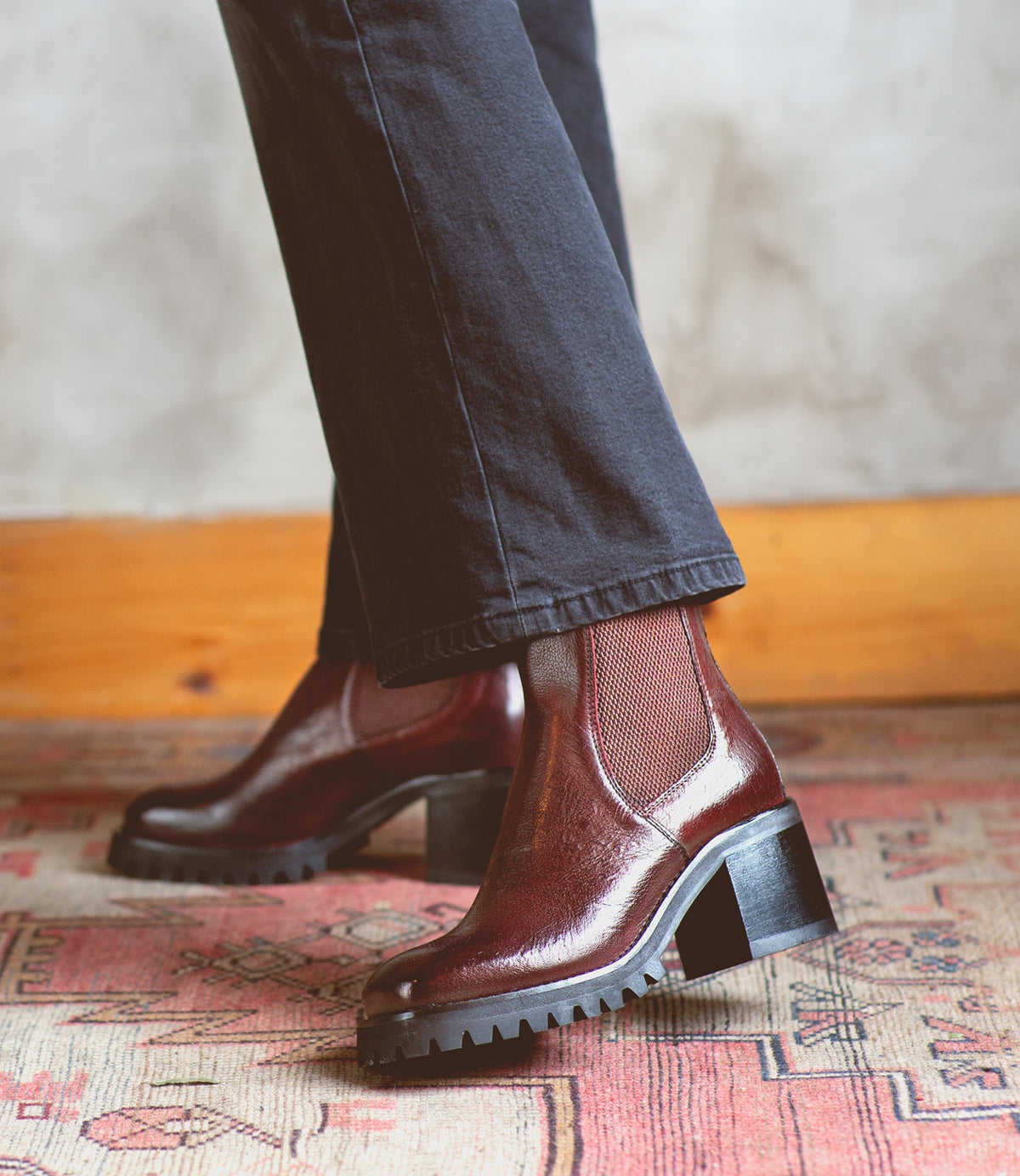 A person wearing black pants stands on a patterned rug, showcasing the Surge dark brown Italian leather ankle boots with chunky heels and lug soles by Bed|Stü.