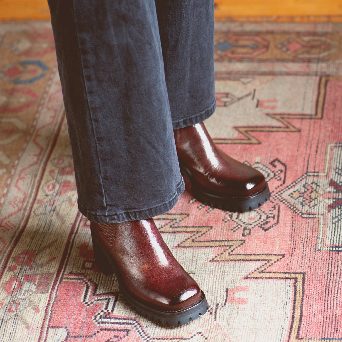Wearing the Surge ankle boots in burgundy Italian leather by Bed|Stü, a person dressed in dark pants stands on a rug with geometric patterns.