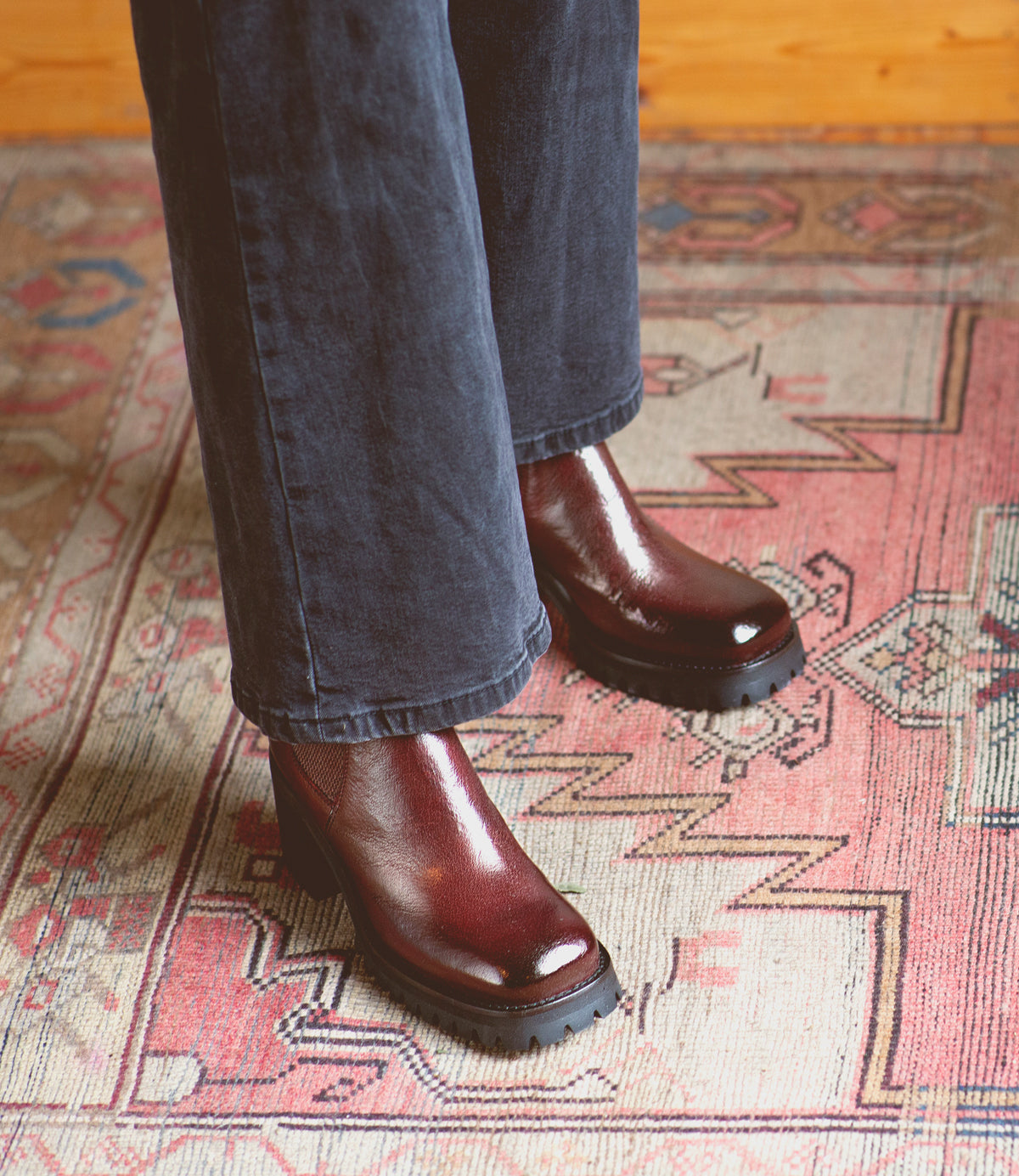 Wearing the Surge ankle boots in burgundy Italian leather by Bed|Stü, a person dressed in dark pants stands on a rug with geometric patterns.