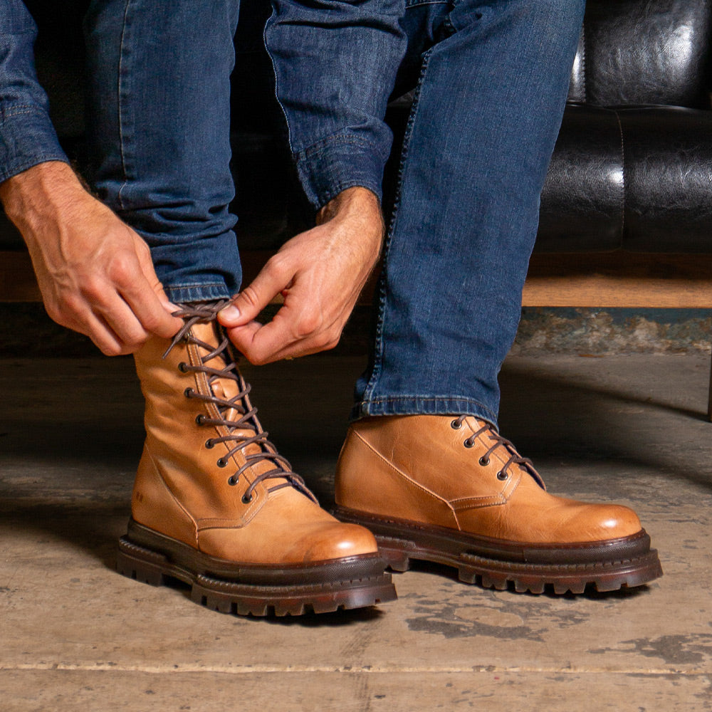 Person tying tan leather lace-up boots with thick treaded soles while wearing blue jeans.