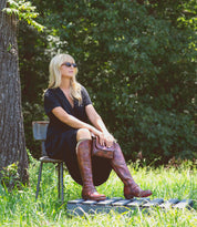 A woman with long blonde hair sits outdoors on a chair, surrounded by trees and grass. She wears sunglasses, a black dress, and the Bed|Stü Takoma tall brown boots featuring a square toe and western-shaped heel, her hands resting on her knees.